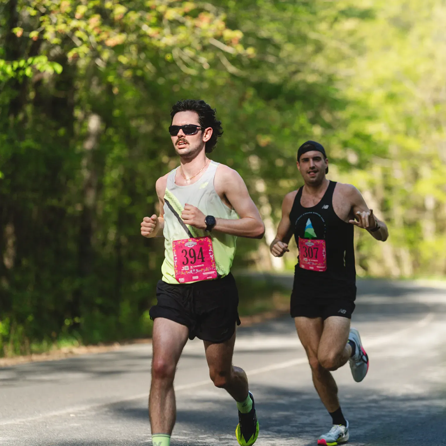 Two male runners on a road course, one in the foreground wearing sunglasses and a light-colored tank top, the other behind him in a dark tank top.
