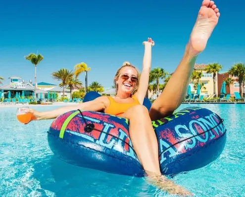 A woman in an orange swimsuit smiles and laughs while floating on a blue tube in a pool, holding a drink.