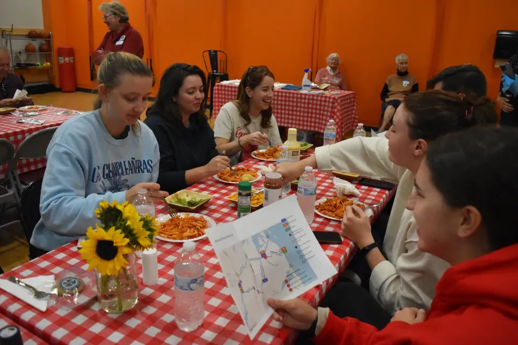 A group of people sitting at a table with red and white checkered tablecloths, eating pasta and smiling, with a map on the table.