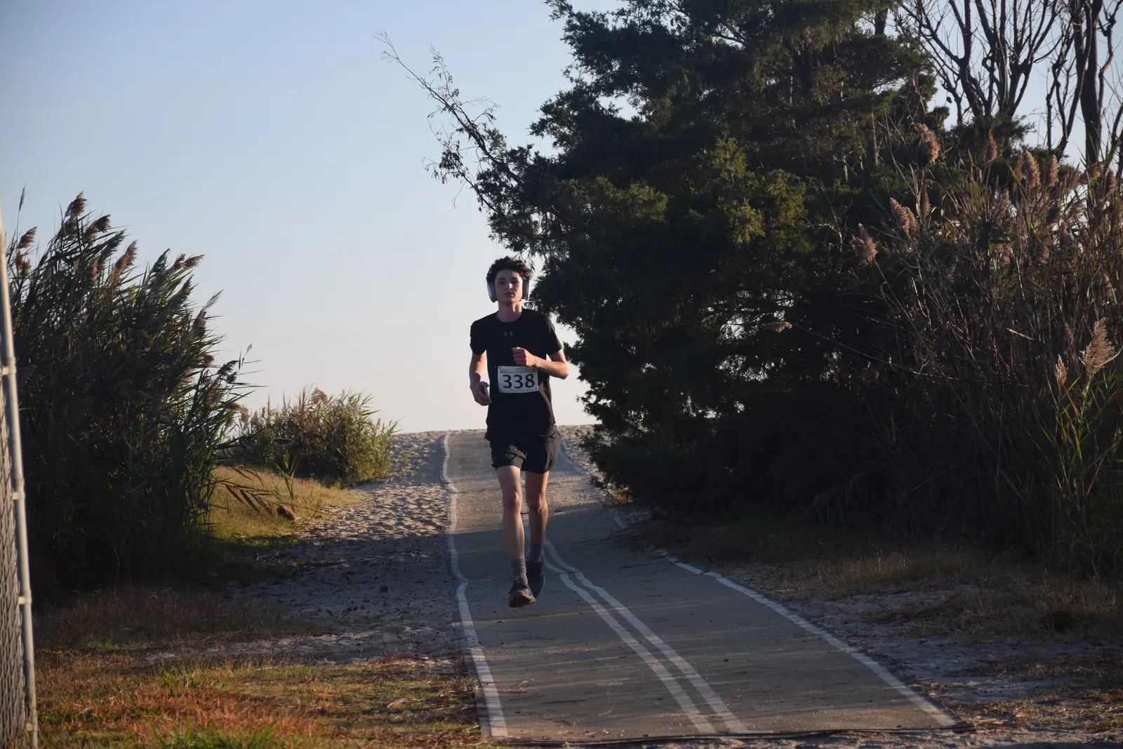 A young male runner wearing headphones and a bib with the number 338 runs on a paved path lined with vegetation.