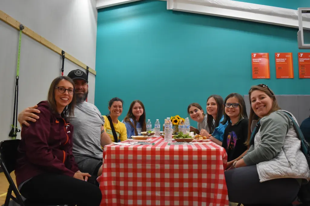 A group of people, including adults and children, smiling and sitting around a table with a red and white checkered tablecloth.