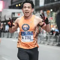 A male runner with bib number A 7781 smiles and holds up two fingers on each hand while running.