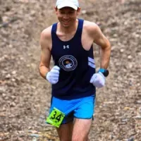 A male runner wearing a navy singlet and blue shorts smiles as he runs on a dirt path, wearing a white cap and white gloves.