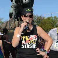 A female runner wearing a black tank top with 'MDMP STRONG' and a Maryland flag design, sunglasses, and a black baseball cap, smiles while holding a water bottle.