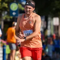 A male runner wearing an orange tank top and red shorts, with a black headband, runs on a road during a race.