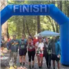 A group of six runners smile for a photo under a blue inflatable finish line archway in a wooded area.