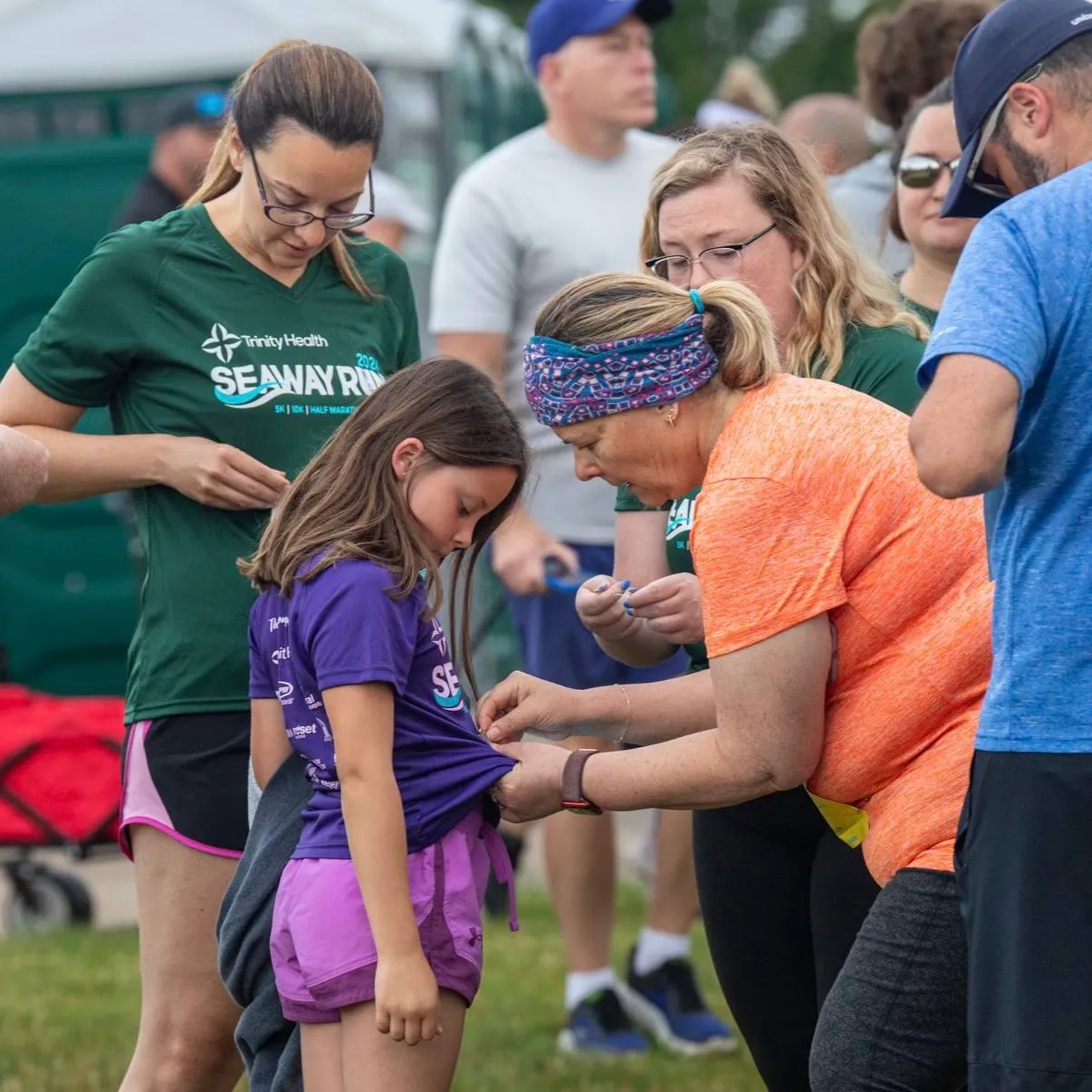 A group of people, including a young girl and adults, are seen preparing for a race, with some adjusting race bibs or shirts. The 'Seaway Run' logo is visible on some of the participants' attire.