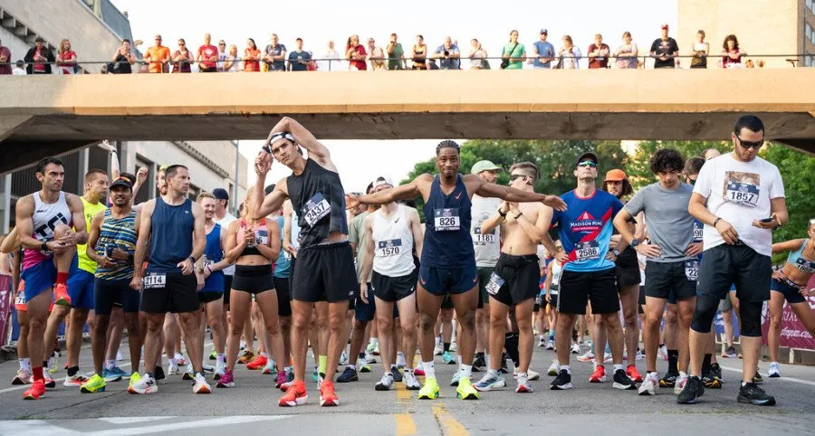 Runners stretching and waiting at the start line of a race, with spectators watching from an overpass.