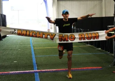 A male runner in a blue cap and black Asics shirt crosses the finish line, breaking a banner that reads 'The Mighty Mo Run'.