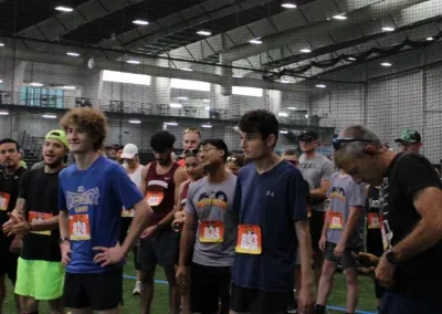 A group of runners, mostly young men, stand in line wearing bib numbers, appearing to wait for a race to begin indoors.
