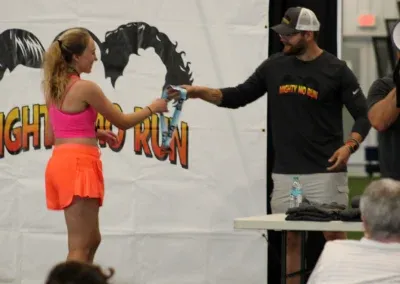 A female runner in a pink top and orange shorts receives a medal from a race official.