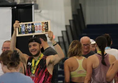 A smiling runner holds up a sign made of license plates that says 'THE STOOGE' and wears a medal around his neck.