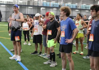 A group of runners in athletic wear, some wearing bib numbers, stand on an indoor track, appearing to be waiting for a race to begin.