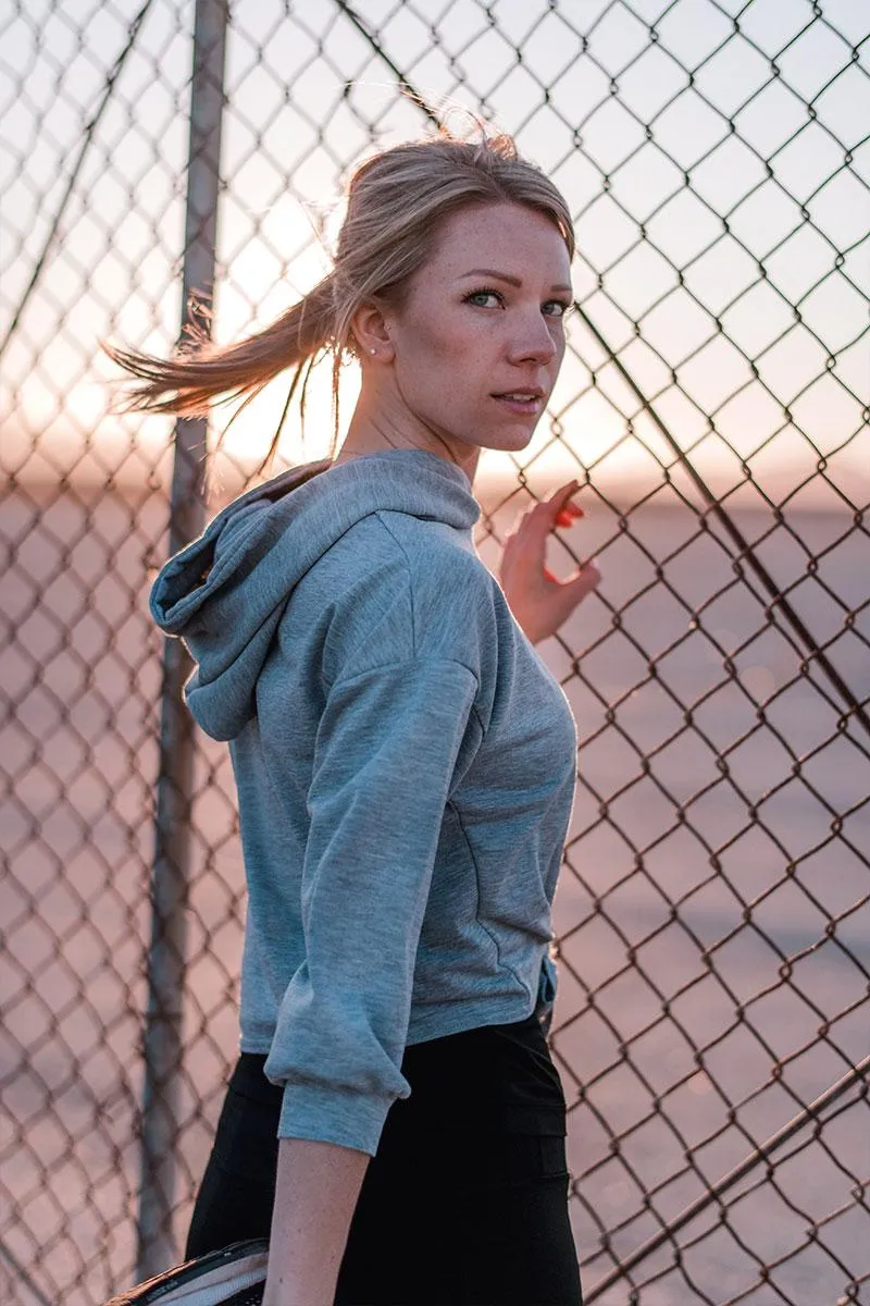 A woman with freckles and her hair in a ponytail, wearing a grey hoodie and black shorts, leans against a chain-link fence at sunset.
