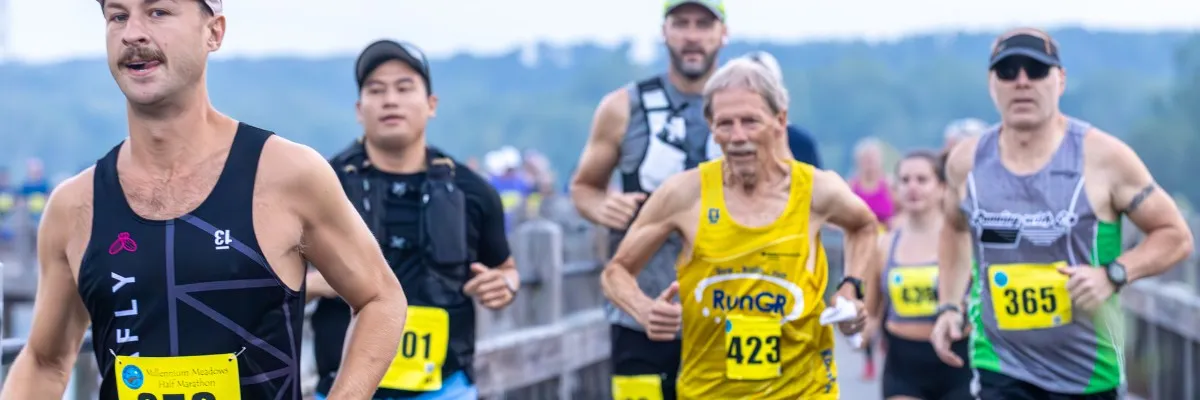 Runners in a race, with the lead runner in a black tank top and a prominent mustache, followed by others in various running attire.