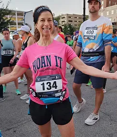 A smiling female runner wearing a pink t-shirt with 'HOAN IT' printed on it, standing at the start line of a race with other runners in the background.