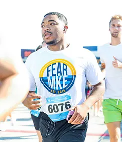 A male runner wearing a white t-shirt with 'FEAR MIKE EST. 2015' and bib number 6076, running in a race.