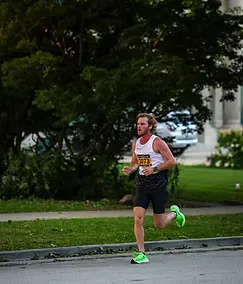 A male runner with a beard runs on a road course, wearing a white tank top and black shorts.