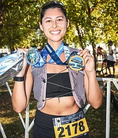 A smiling female runner holds up two race medals, one on a blue ribbon and another on a black ribbon, with a race bib number 2178 visible on her waist.