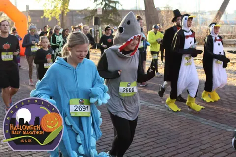 Runners in Halloween costumes, including a shark, a dinosaur, and penguins, participating in the Minneapolis Halloween Half Marathon.
