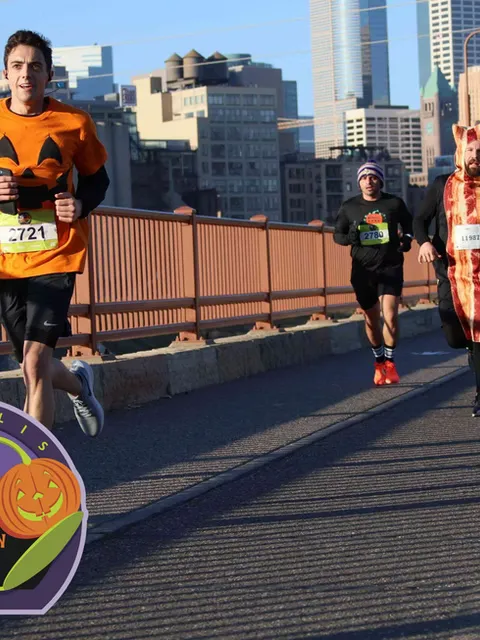 Runners in Halloween costumes, including a pumpkin and a bacon strip, race across a bridge with city buildings in the background.