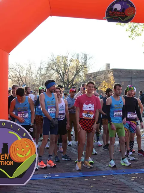 Runners in athletic wear stand at the start line of the Minneapolis Halloween Half Marathon, with an orange inflatable arch overhead and a pumpkin logo visible.