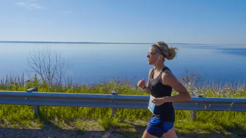 A female runner with headphones runs along a road with a large body of water in the background.
