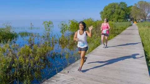 A woman smiles and waves as she runs on a wooden boardwalk next to a body of water, with another runner behind her.