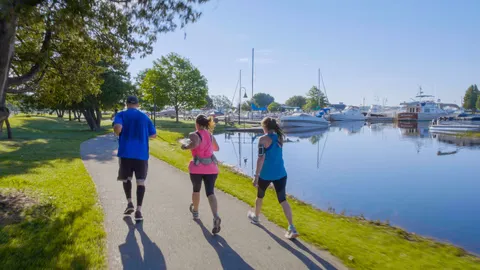Three runners jogging on a paved path next to a calm body of water with sailboats docked in the background.