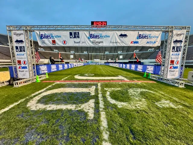 The start/finish line of the Mississippi Blues Marathon, set up on a football field with banners and a clock displaying the time.