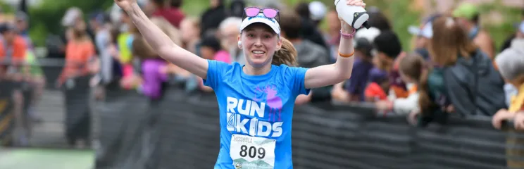 A female runner with her arms raised in celebration, wearing a blue 'RUN FOR KIDS' shirt and a white visor, crosses the finish line with a crowd watching in the background.