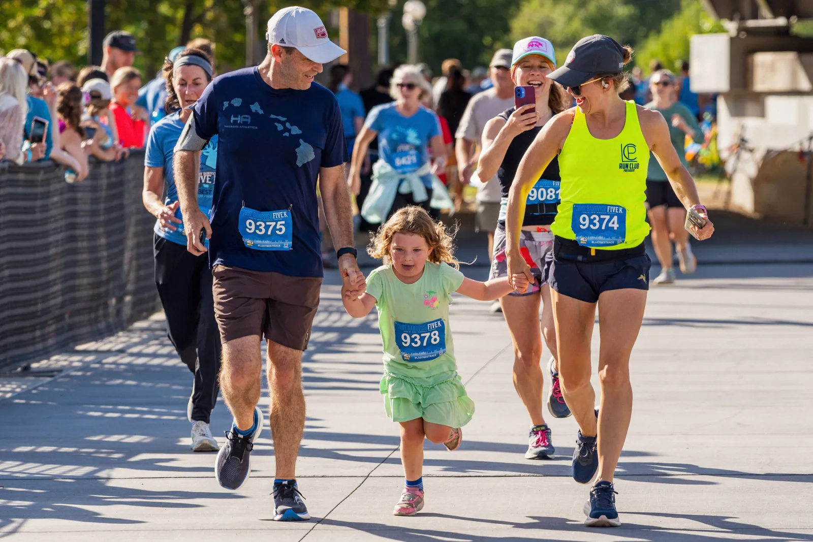 A man and a young girl hold hands while running in a race, with other runners and spectators in the background.