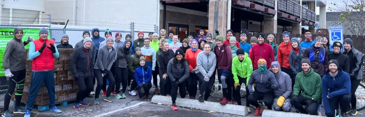 A large group of runners, dressed in athletic wear and hats, pose for a photo outdoors in a parking lot.