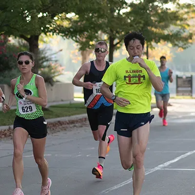 Three runners on a paved course, with the lead runner in a bright yellow shirt and the runner to their left in a green tank top with bib number 629.