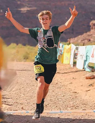 A young male runner with bib number 772 crosses the finish line with arms raised in celebration.