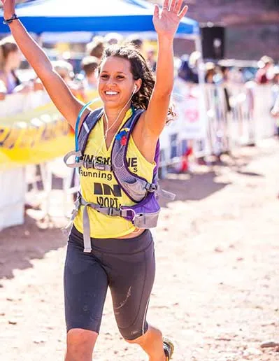 A female runner with her arms raised in celebration as she crosses the finish line, wearing a yellow shirt and dark shorts.