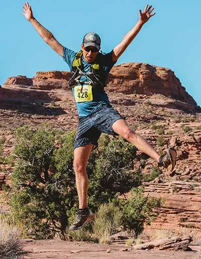A runner jumps in the air with arms raised in celebration on a desert trail.