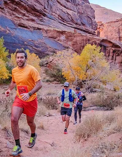Three runners on a dusty trail with red rock cliffs and autumn foliage in the background. The lead runner in an orange shirt is smiling at the camera.