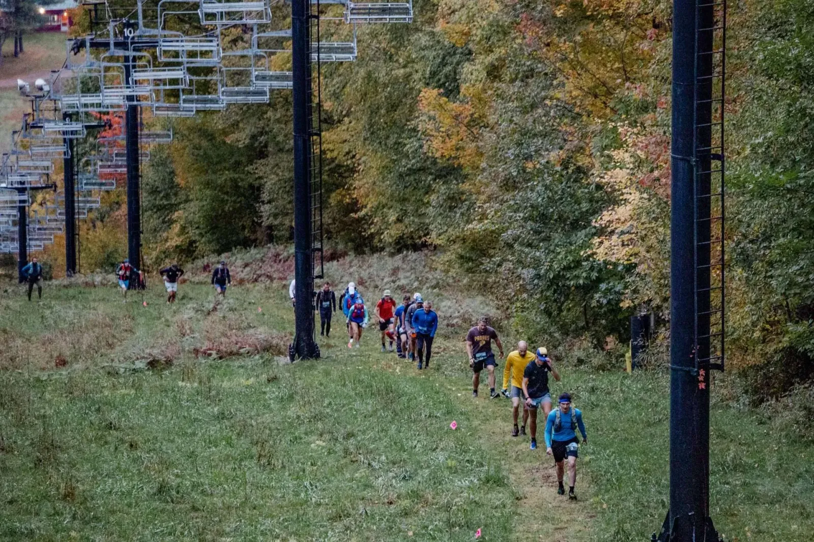 Runners ascending a grassy slope with ski lifts and autumn foliage in the background.