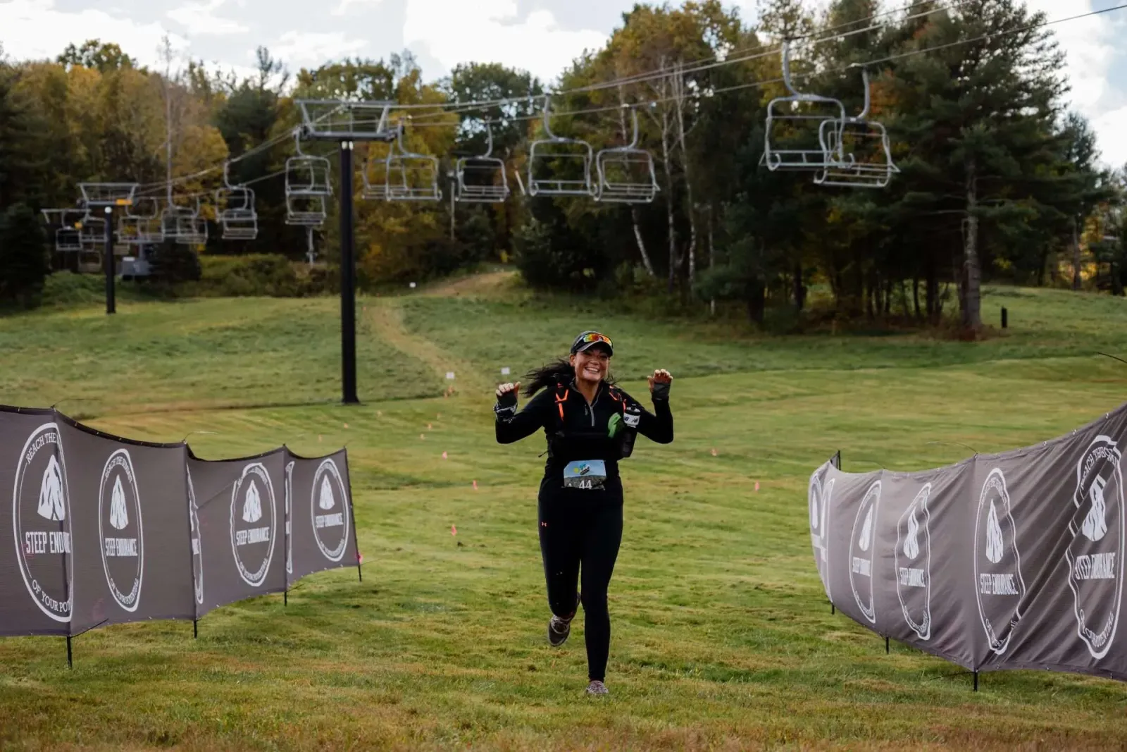 A female runner with her arms raised in celebration runs towards the camera on a grassy slope, with ski lifts visible in the background and race banners lining the course.