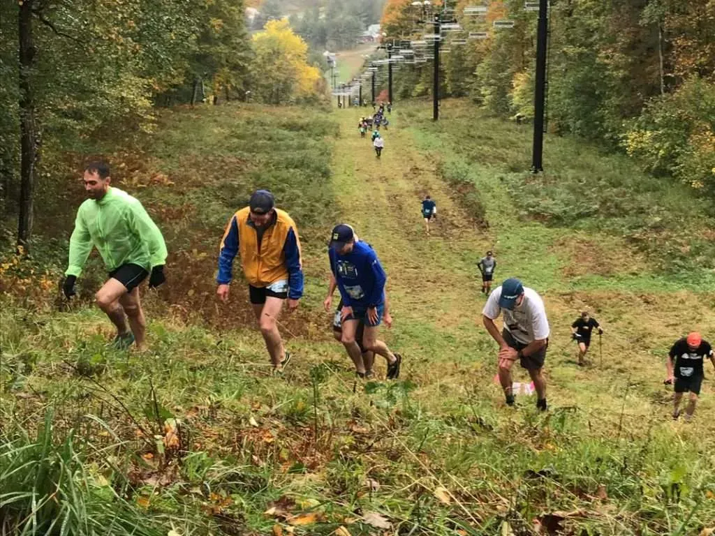 Runners ascending a steep, grassy slope with ski lifts in the background during a mountain race.