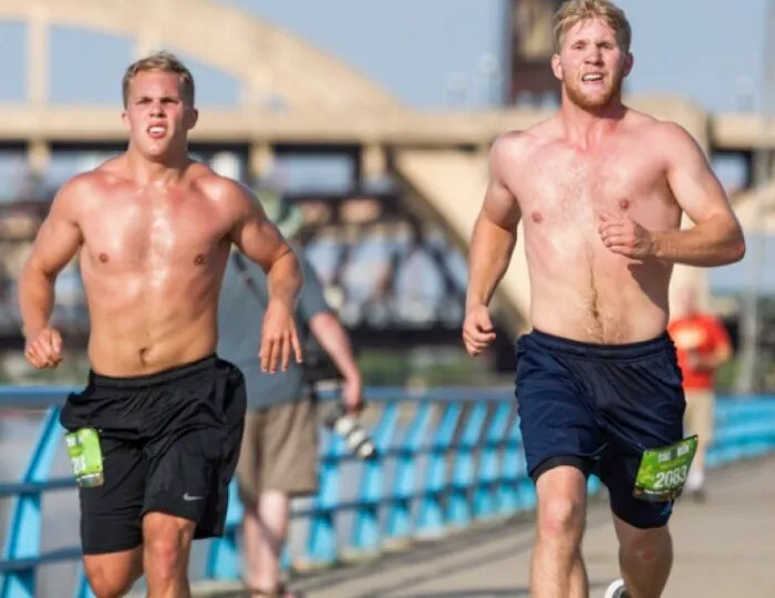 Two shirtless male runners with bib numbers on their shorts running on a bridge.