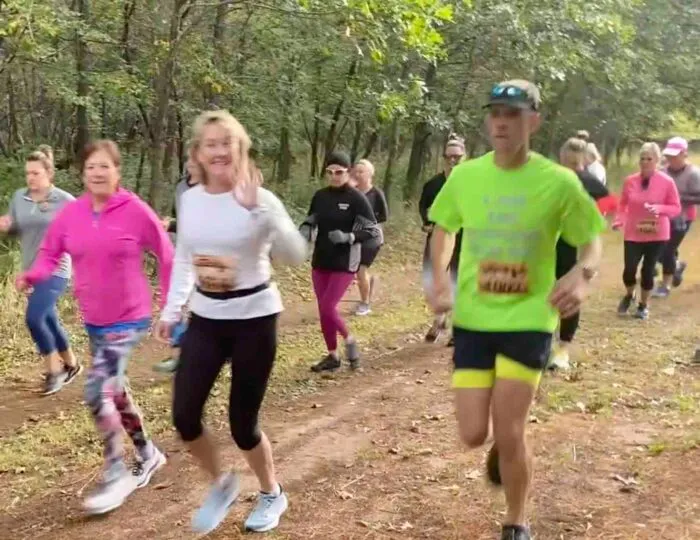 A group of runners on a wooded trail, with some smiling and waving.