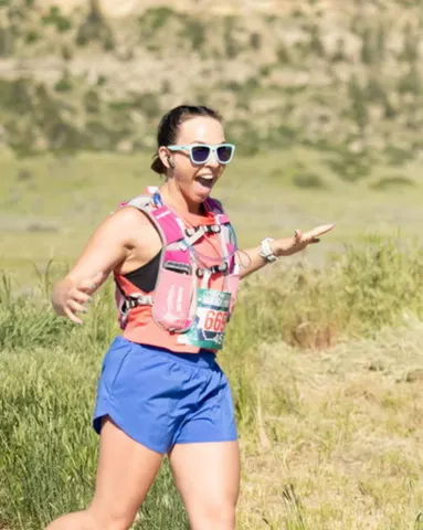 A female runner with a look of excitement on her face, wearing sunglasses and a hydration pack, running on a grassy course with hills in the background.