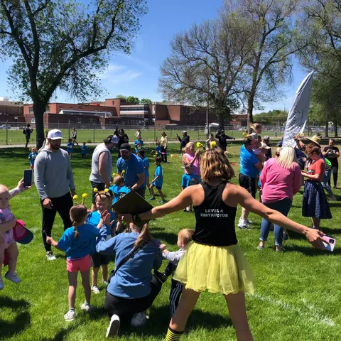 A group of people, including children and adults, gathered on a grassy field under a sunny sky, with some children wearing bee-like antennae. An instructor in the foreground is facing away from the camera, wearing a black tank top and a yellow skirt.