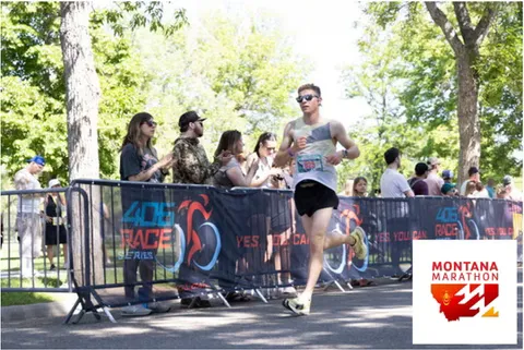 A male runner in sunglasses and a patterned tank top runs past spectators behind a barrier at the Montana Marathon.