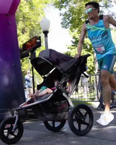 A runner pushes a baby in a stroller past a timer at the start line of a race.