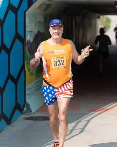 A male runner wearing an orange race bib and patriotic shorts gives a thumbs up while running through a tunnel with colorful murals.