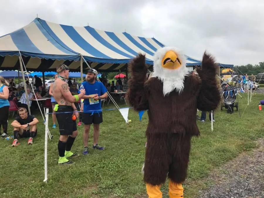 A person in an eagle mascot costume with arms raised, standing in front of a tent at a race event. Two runners are talking in the background.