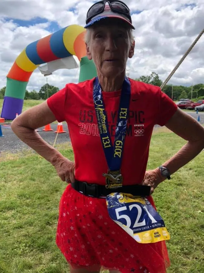An older female runner stands proudly wearing a red shirt, a race medal, and a bib with the number 521. She is in front of a colorful inflatable archway, suggesting she has just finished a race.